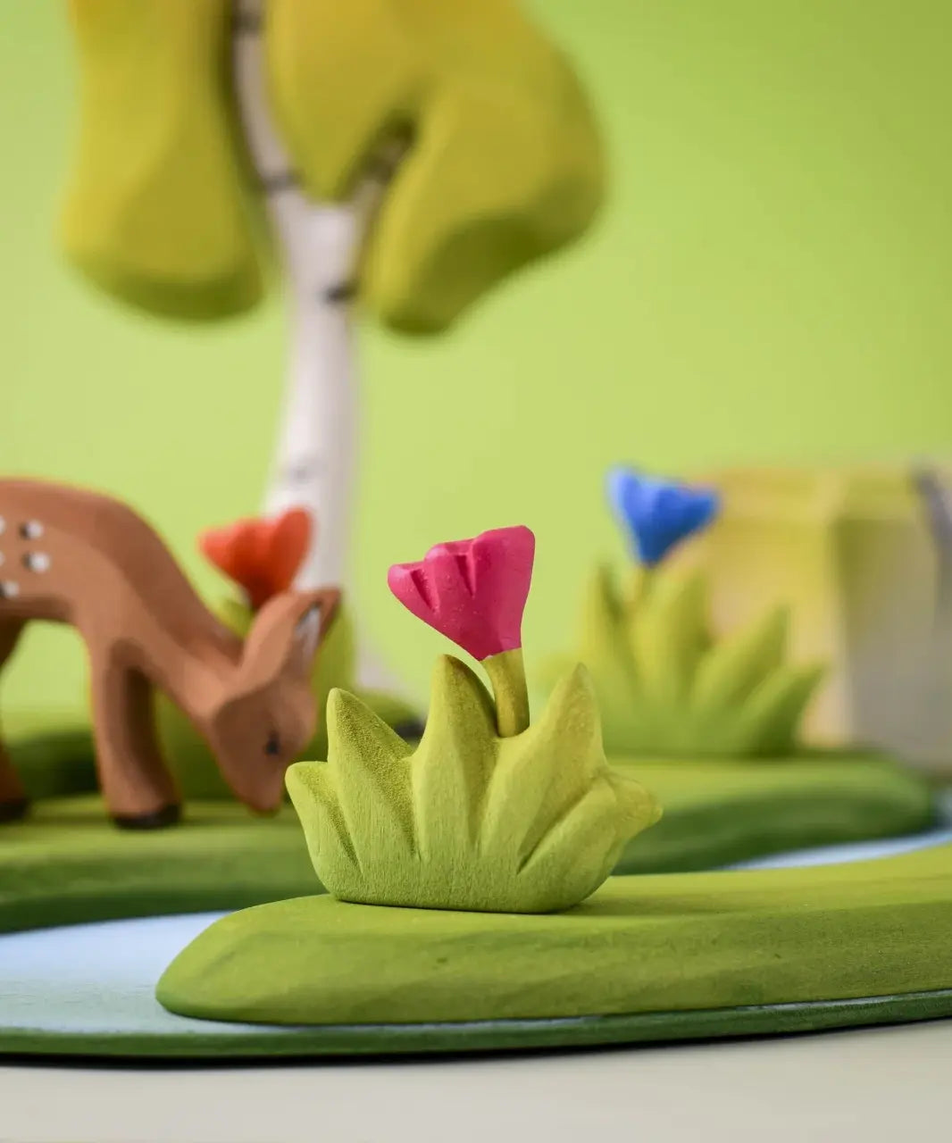 Bumbu small wooden grass with pink flower placed at the front of a small world play scene with deer, flowers and trees in the background.