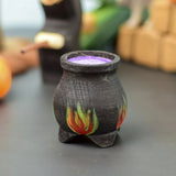 A Bumbu Wooden Witch's Cauldron. The cauldron is black on the outside and decorated with red, yellow and orange painted flames on the side. The top of the cauldron has a purple swirled effect decoration. The cauldron is placed at the front of a Halloween themed play scene, other wooden toys can be seen in the background. 