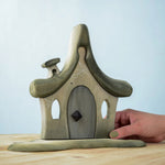 Bumbu Toys Witches house decorated with a Halloween theme. The house is grey in colour hand has cobwebs and a spider painted on. The House is placed on a wooden surface and an adult's hand can be seen holding one side. 