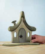 Bumbu Toys Witches house decorated with a Halloween theme. The house is grey in colour hand has cobwebs and a spider painted on. The House is placed on a wooden surface and an adult's hand can be seen holding one side. 