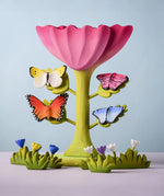 Bumbu Toys wooden Butterflies placed on a Bumbu Pink Flower, surrounded by Bumbu flowers and grass.