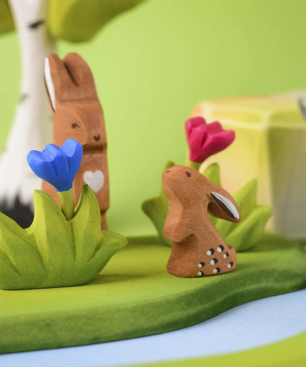 The Bumbu Toys wooden baby rabbit, in a curious upright position and surrounded by Bumbu wooden creatures and nature