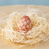 A Bumbu wooden egg placed in a wooden wool nest on a wooden surface. 