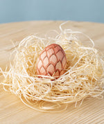 A Bumbu wooden egg placed in a wooden wool nest on a wooden surface. 