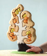 The Bumbu Toys Big Puzzle Birch Tree showing the orange and yellow leaves side placed on a wooden surface in front of a blue background with an adult's hand holding on to the trunk.