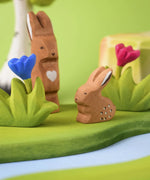 The Bumbu Toys wooden baby rabbit figure, in a sitting position and surrounded by Bumbu landscape and nature toys