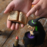 A close up of a  Bumbu Toys Open Spell Book in a child's hand. The hand is pointing at the markings in the book. Part of Bumbu's Handmade Halloween Collection. These expertly hand crafted items are part of a large range of wooden toys available here at Babipur. 