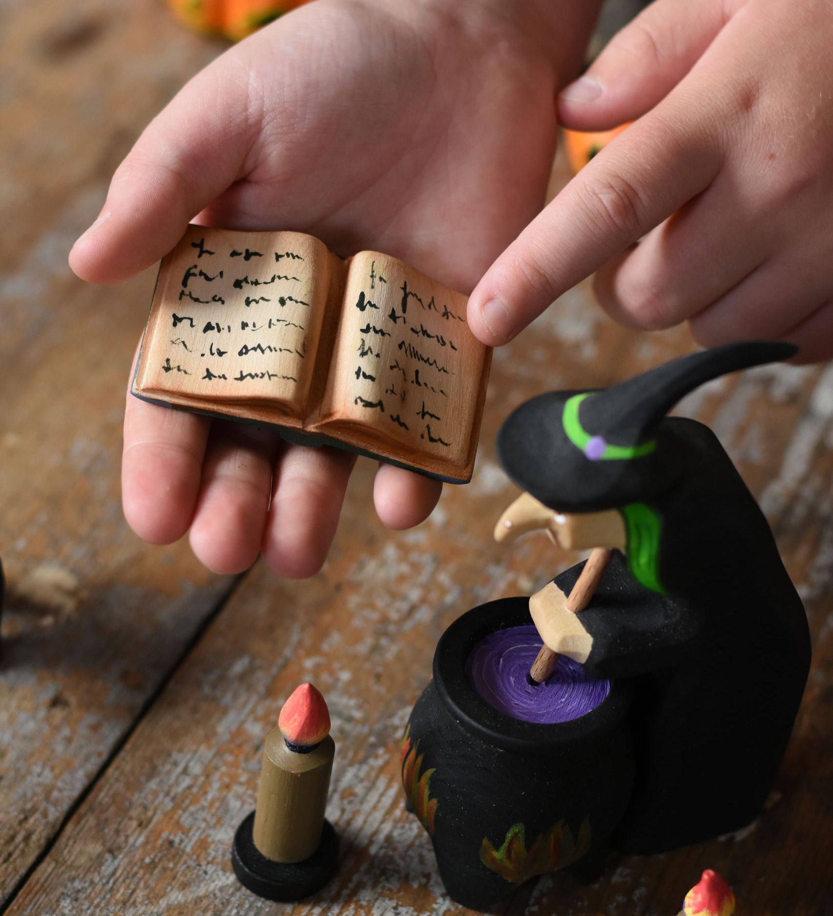 A close up of a  Bumbu Toys Open Spell Book in a child's hand. The hand is pointing at the markings in the book. Part of Bumbu's Handmade Halloween Collection. These expertly hand crafted items are part of a large range of wooden toys available here at Babipur. 