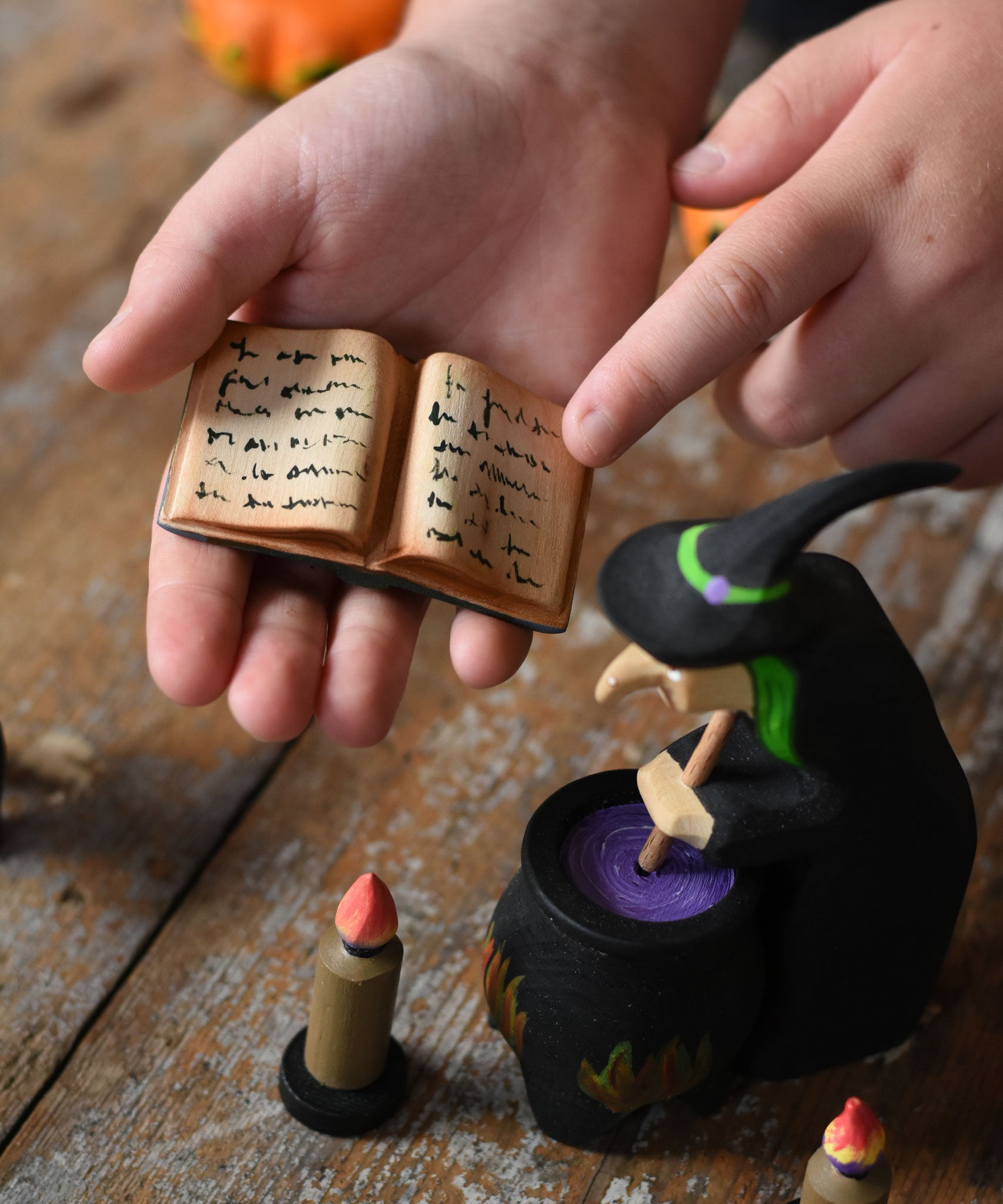 A close up of a  Bumbu Toys Open Spell Book in a child's hand. The hand is pointing at the markings in the book. Part of Bumbu's Handmade Halloween Collection. These expertly hand crafted items are part of a large range of wooden toys available here at Babipur. 