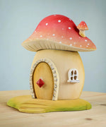 The Bumbu Toys Toadstool House placed on a wooden surface. The toadstool roof is red with white dots and has a matching small toadstool as a chimney.  There is a little white sash window and a door with a red diamond knob in the centre