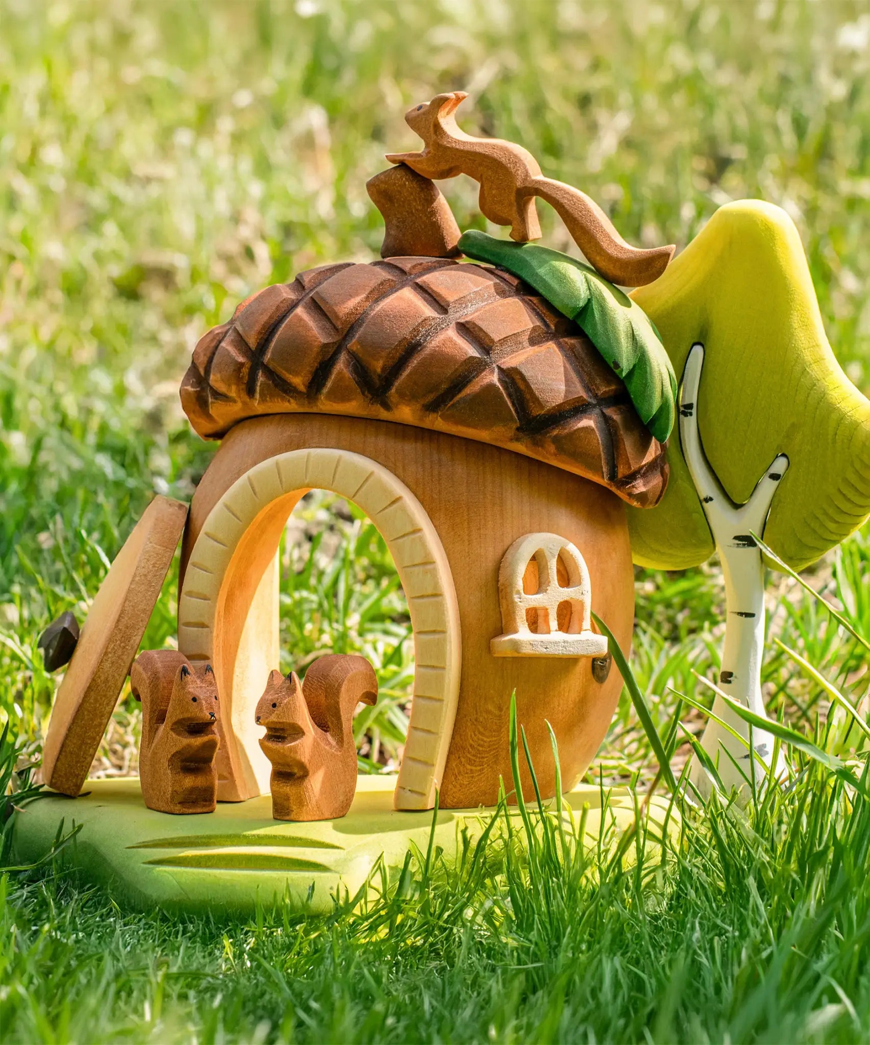 Bumbu toys handcrafted wooden acorn house with the removable door open, squirrel figures on the base and tree figure behind, standing in a grassy field