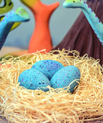 A set of Bumbu Wooden Brontosaurus Dinosaur Eggs in a nest. Dinosaurs and wooden trees can be seen in the background. 