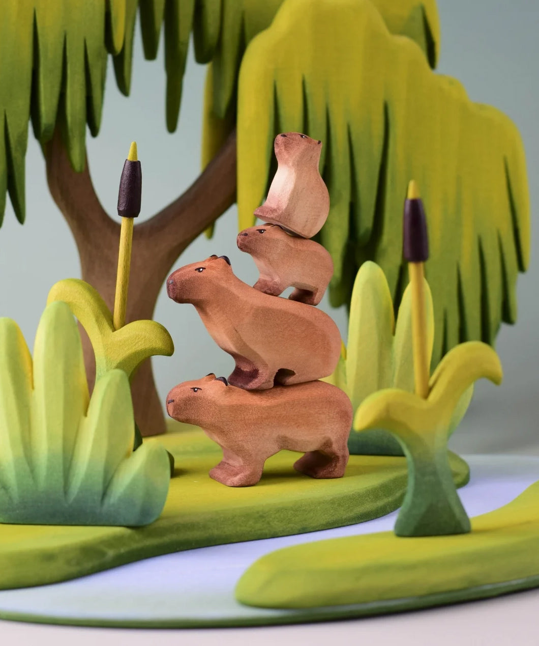 Bumbu Toys Wooden Capybara, Baby Capybara, Sitting Capybara and Baby Sitting Capybara, all stacked on top of each other. They are surrounded by Bumbu trees and grass.
