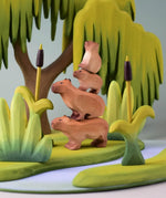 Bumbu Toys Wooden Capybara, Baby Capybara, Sitting Capybara and Baby Sitting Capybara, all stacked on top of each other. They are surrounded by Bumbu trees and grass.
