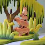 Bumbu Toys Wooden Capybara, Baby Capybara, Sitting Capybara and Baby Sitting Capybara, all stacked on top of each other. They are surrounded by Bumbu trees and grass.