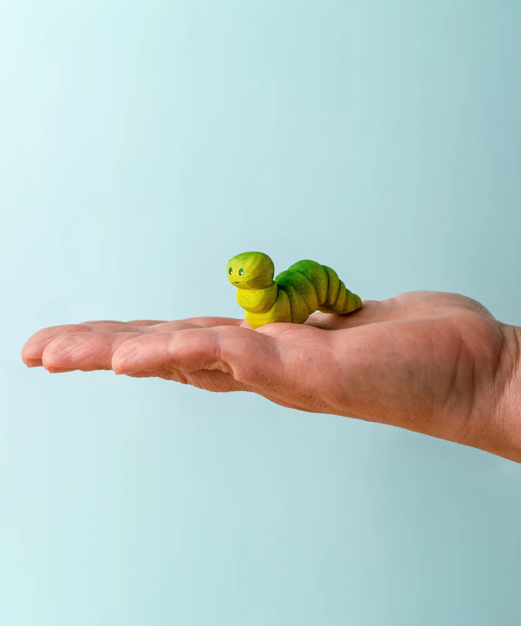 Adult hand holding a Bumbu handcrafted wooden green caterpillar figure showing scale