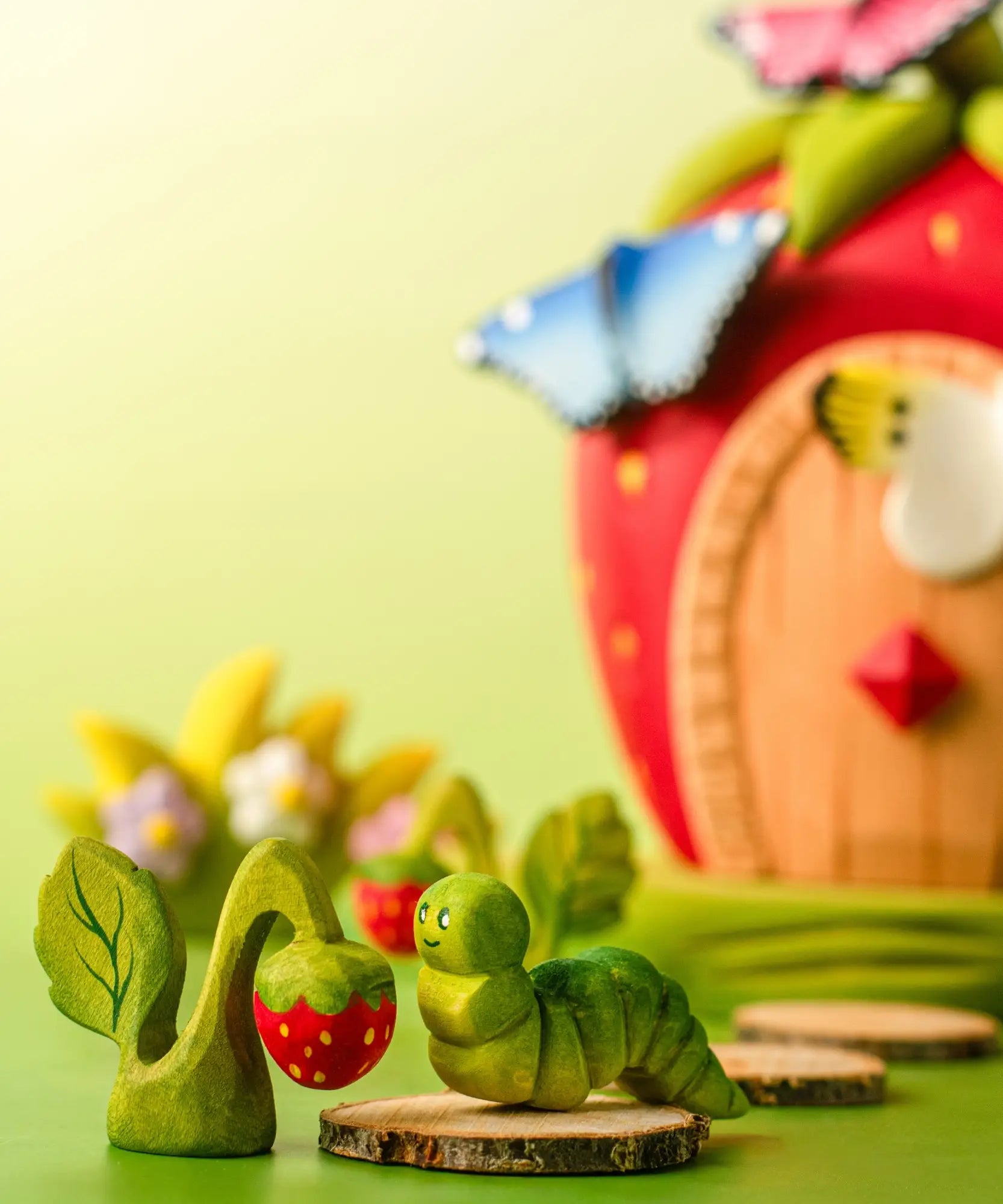 Bumbu handcrafted wooden green caterpillar figure next to a strawberry plant figure and a strawberry house in the backhground.