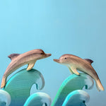 Two Bumbu Wooden Dolphins perched on waves in front of a blue background. 
