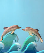 Two Bumbu Wooden Dolphins perched on waves in front of a blue background. 