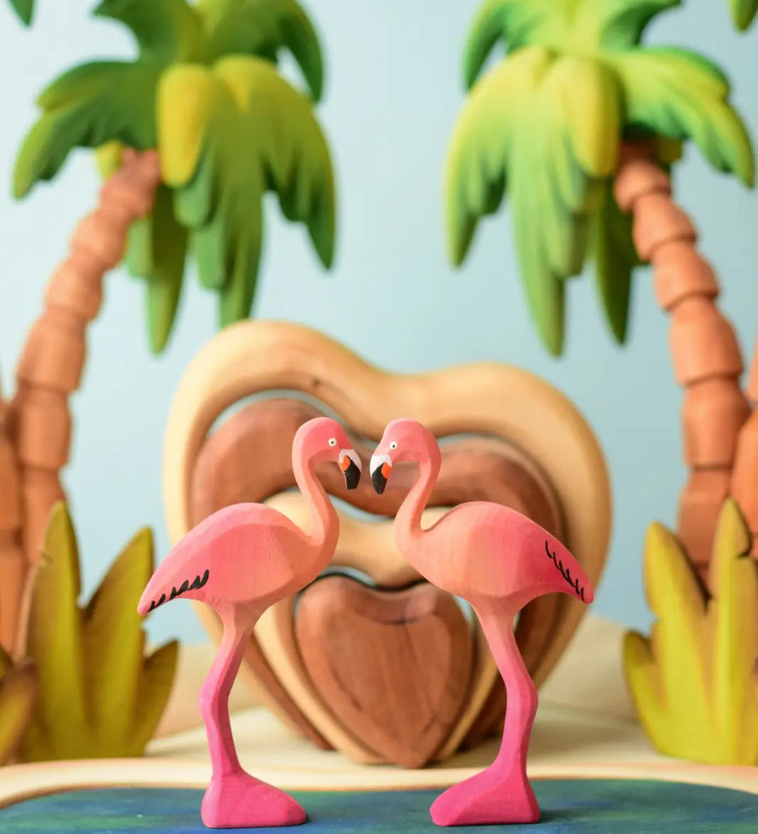 2 Bumbu toys handcrafted wooden flamingo figures side by side with a heart stacker set and palm trees in the background