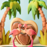 2 Bumbu toys handcrafted wooden flamingo figures side by side with a heart stacker set and palm trees in the background