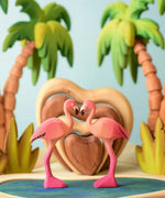 2 Bumbu toys handcrafted wooden flamingo figures side by side with a heart stacker set and palm trees in the background