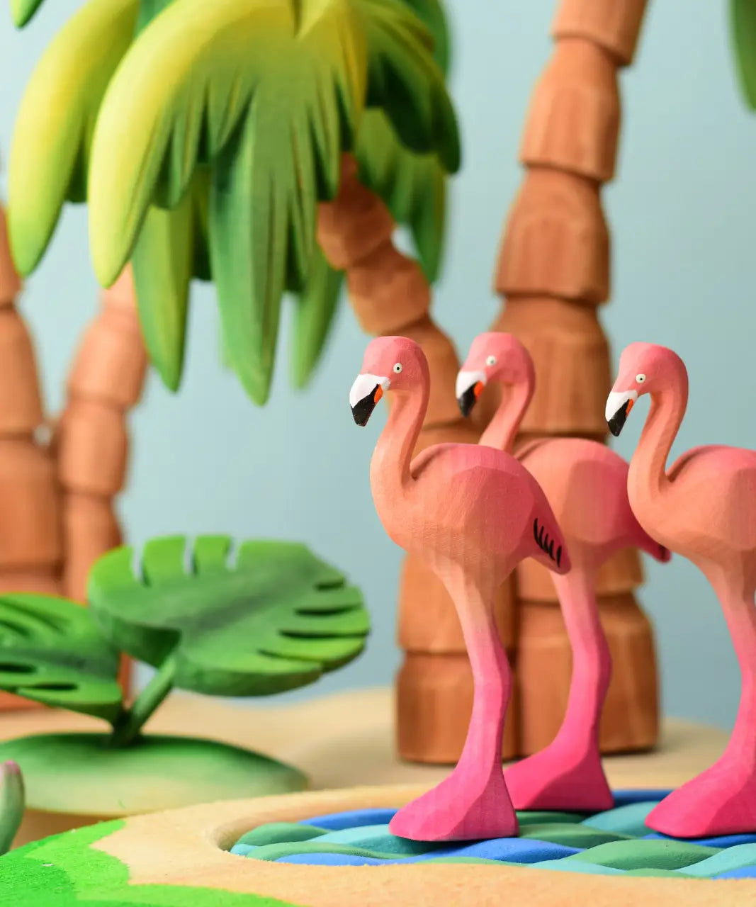 Play scene showing Bumbu toys handcrafted wooden flamingo figures on a pond and palm tree figures in the background