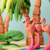 Play scene showing Bumbu toys handcrafted wooden flamingo figures on a pond and palm tree figures in the background