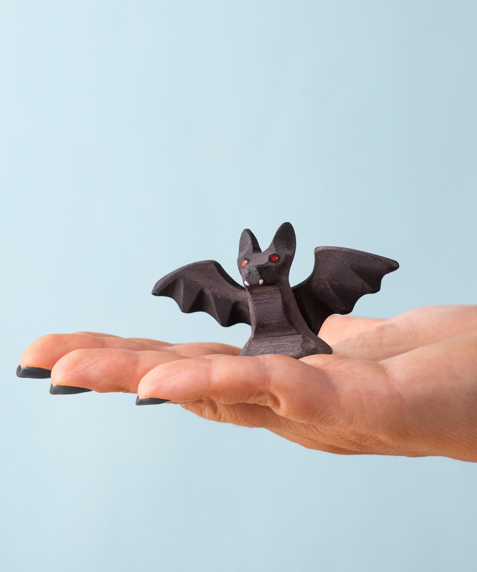 An adult hand holding the Bumbu handmade and hand painted wooden standing bat figure available at Babipur showing its size