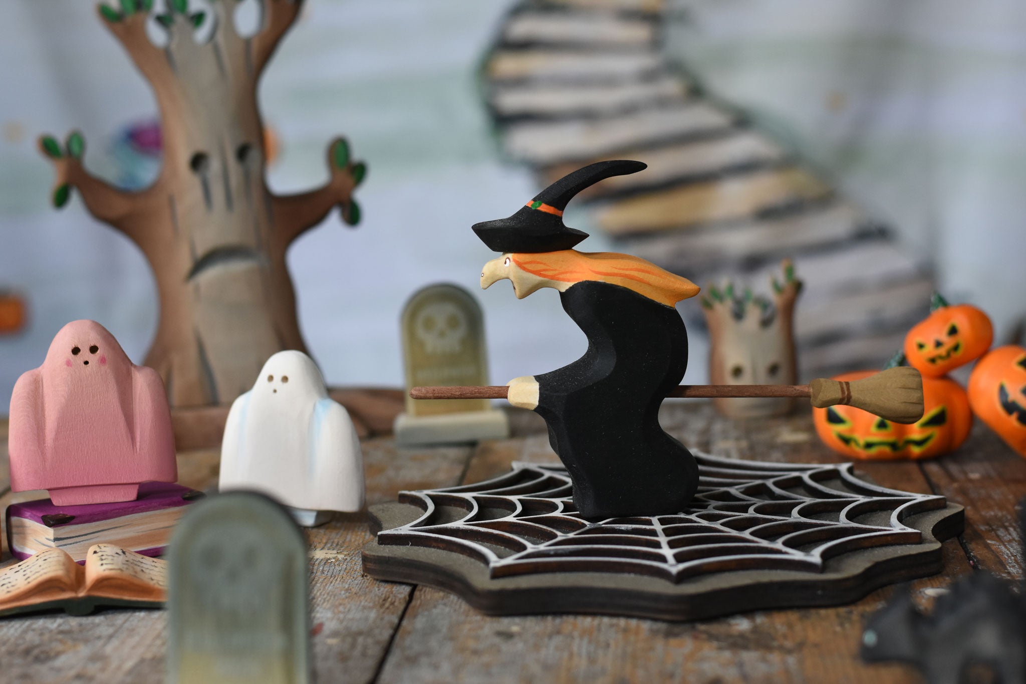 A Bumbu witch on a broom figure placed in the middle of a Halloween themed small world play scene. The witch has been placed on a wooden cobweb and is surrounded by ghosts, spell books, bumpkins, gravestones and a spooky tree in the background. These expertly hand crafted items are part of a large range of wooden toys available here at Babipur. 
