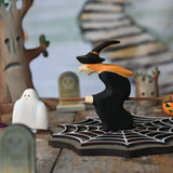 A Bumbu witch on a broom figure placed in the middle of a Halloween themed small world play scene. The witch has been placed on a wooden cobweb and is surrounded by ghosts, spell books, bumpkins, gravestones and a spooky tree in the background. These expertly hand crafted items are part of a large range of wooden toys available here at Babipur. 