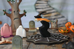A Bumbu witch on a broom figure placed in the middle of a Halloween themed small world play scene. The witch has been placed on a wooden cobweb and is surrounded by ghosts, spell books, bumpkins, gravestones and a spooky tree in the background. These expertly hand crafted items are part of a large range of wooden toys available here at Babipur. 