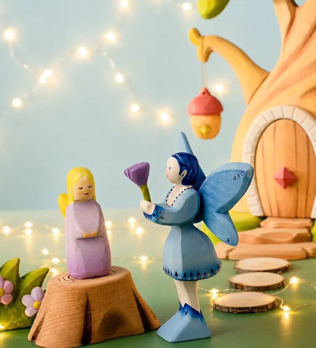 A Bumbu wooden lavender fairy figure standing on a tree stump next to a blue fairy and a treehouse in the background.