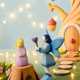 A Bumbu wooden lavender fairy figure standing on a tree stump next to a blue fairy and a treehouse in the background.