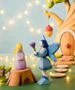 A Bumbu wooden lavender fairy figure standing on a tree stump next to a blue fairy and a treehouse in the background.