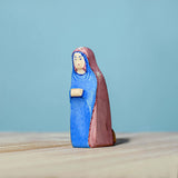 Bumbu Handmade Wooden Mary Figure kneeling down on a wooden table and blue background.