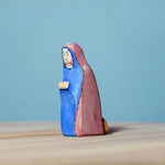 Bumbu Handmade Wooden Mary Figure kneeling down on a wooden table and blue background.