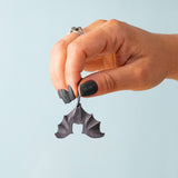 An adult hand holding the Bumbu handmade and hand painted wooden hanging bat figure available at Babipur showing its size