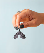 An adult hand holding the Bumbu handmade and hand painted wooden hanging bat figure available at Babipur showing its size