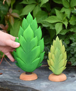 A hand reaching out for the large thuja wooden tree that has been placed on a slate surface outdoors. There is a small thuja tree next to it. These expertly hand crafted items are part of a large range of wooden toys available here at Babipur. 