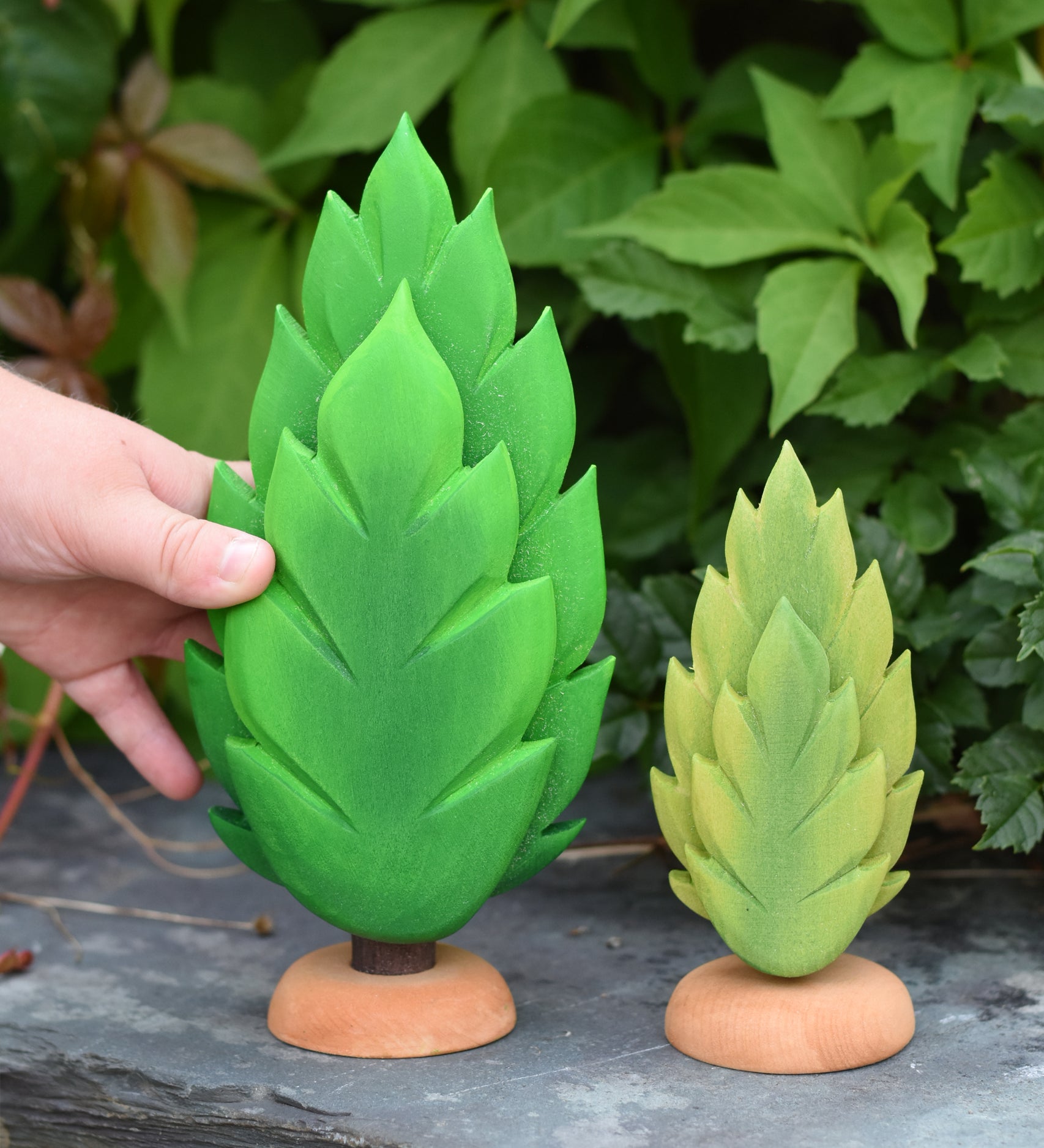 A hand reaching out for the large thuja wooden tree that has been placed on a slate surface outdoors. There is a small thuja tree next to it. These expertly hand crafted items are part of a large range of wooden toys available here at Babipur. 