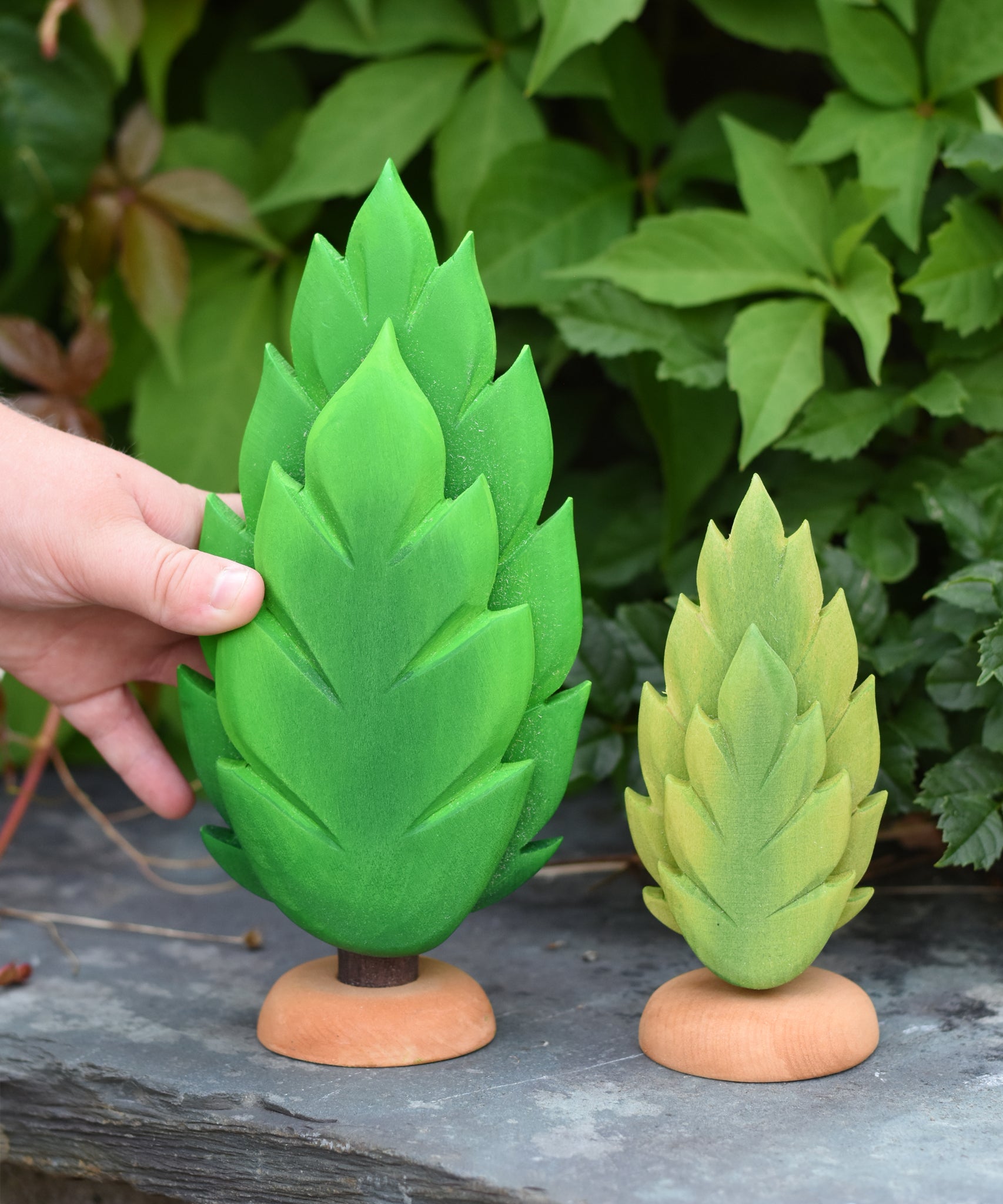 A hand reaching out for the large thuja wooden tree that has been placed on a slate surface outdoors. There is a small thuja tree next to it. These expertly hand crafted items are part of a large range of wooden toys available here at Babipur. 