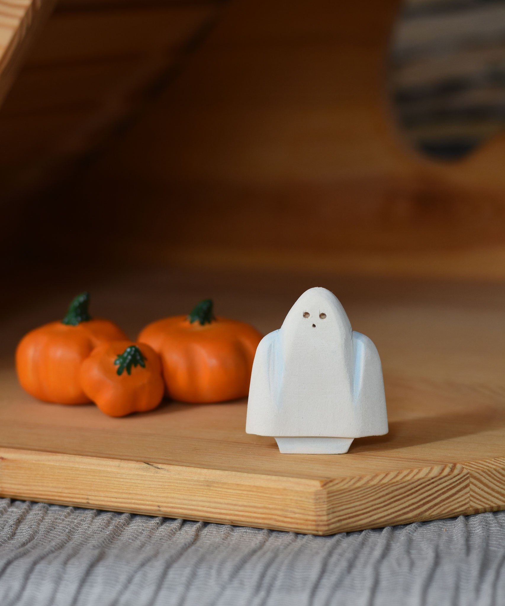 A close up of a Bumbu white ghost figure placed next to three wooden pumpkins in an Ostheimer wooden cottage. 