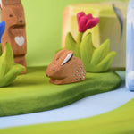 The Bumbu Toys wooden baby brown rabbit, sat in a perching position and surrounded by wooden grass scenery and flowers