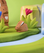 The Bumbu Toys wooden baby brown rabbit, sat in a perching position and surrounded by wooden grass scenery and flowers