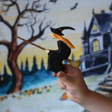 A close up of a Bumbu Witch on broom in a child's hand. the child is holding the witch up in front of a Wonderie play cloth with a Halloween trick or treat design on which has been hung up on a wall. These expertly hand crafted items are part of a large range of wooden toys available here at Babipur. 
