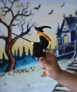 A close up of a Bumbu Witch on broom in a child's hand. the child is holding the witch up in front of a Wonderie play cloth with a Halloween trick or treat design on which has been hung up on a wall. These expertly hand crafted items are part of a large range of wooden toys available here at Babipur. 