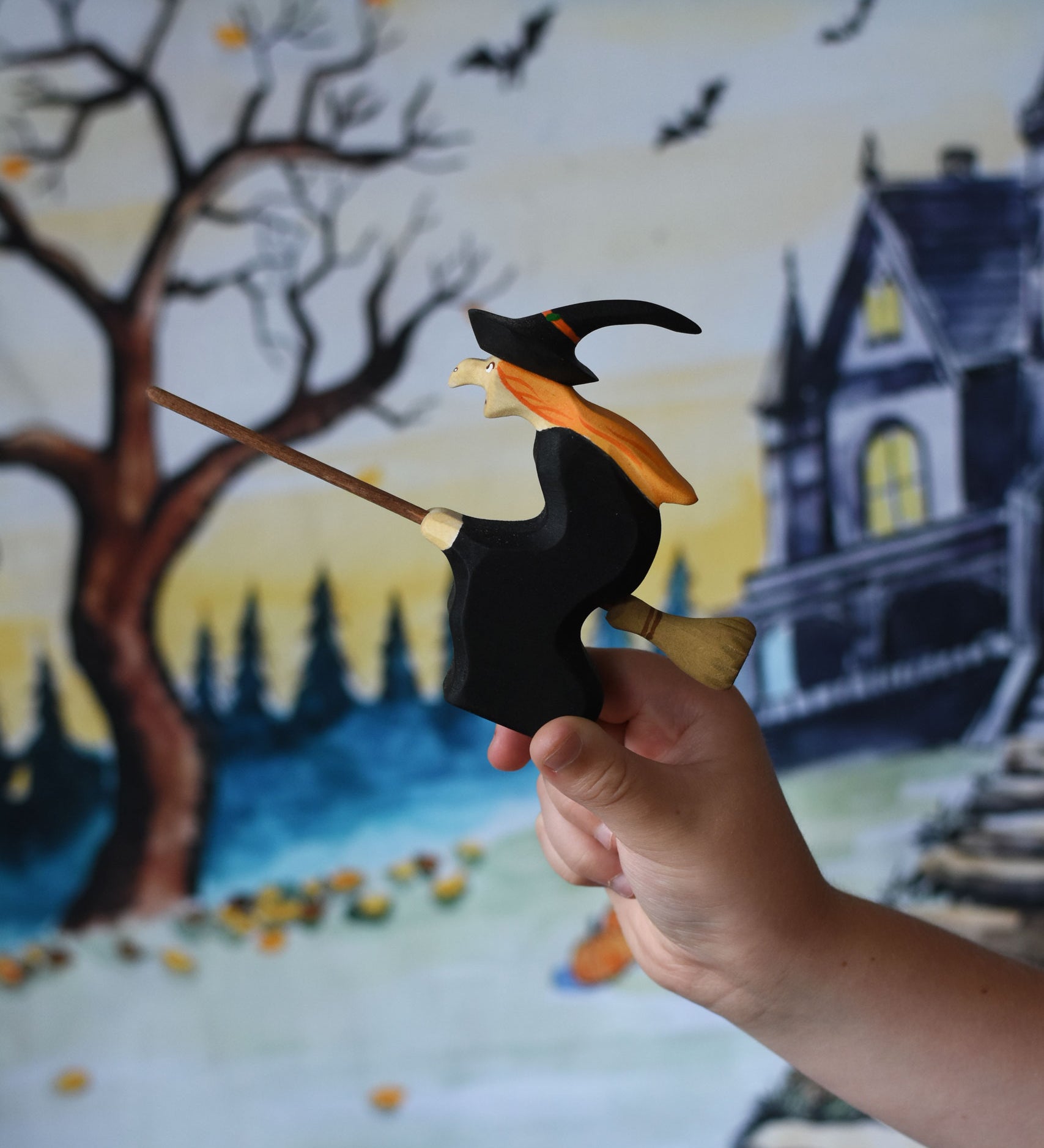 A close up of a Bumbu Witch on broom in a child's hand. the child is holding the witch up in front of a Wonderie play cloth with a Halloween trick or treat design on which has been hung up on a wall. These expertly hand crafted items are part of a large range of wooden toys available here at Babipur. 