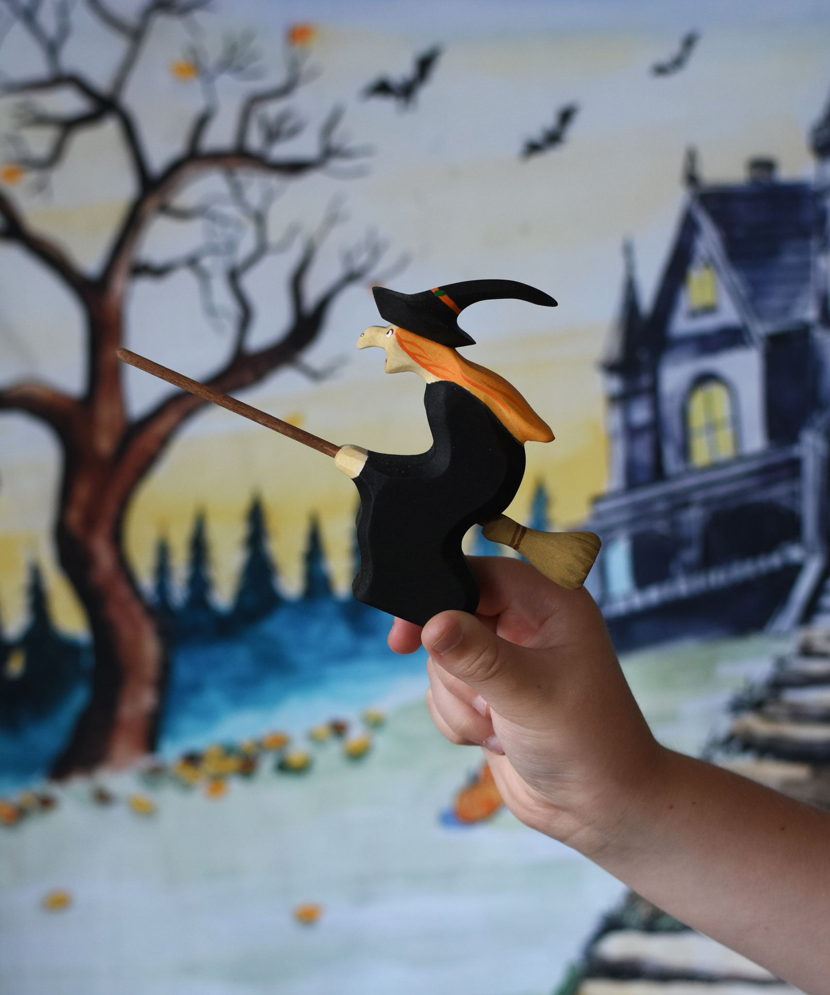 A close up of a Bumbu Witch on broom in a child's hand. the child is holding the witch up in front of a Wonderie play cloth with a Halloween trick or treat design on which has been hung up on a wall. These expertly hand crafted items are part of a large range of wooden toys available here at Babipur. 