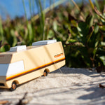 Close up of the Candylab wooden diecast campervan toy on some light sand in front of some green leaves